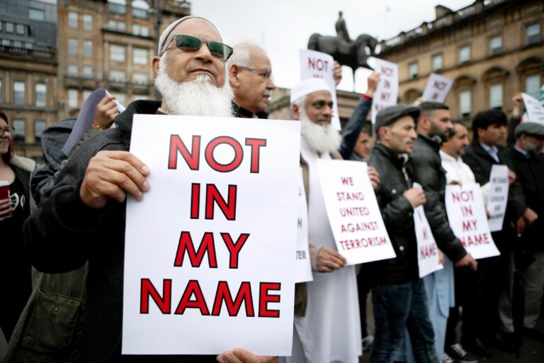 Protesters hold signs reading "Not in My Name" and "We Stand United Against Terrorism" in a city square, conveying unity and peaceful resistance.