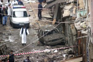 Forensic investigators examine a street littered with debris from a building explosion. A damaged car and police tape are visible, conveying chaos and urgency.