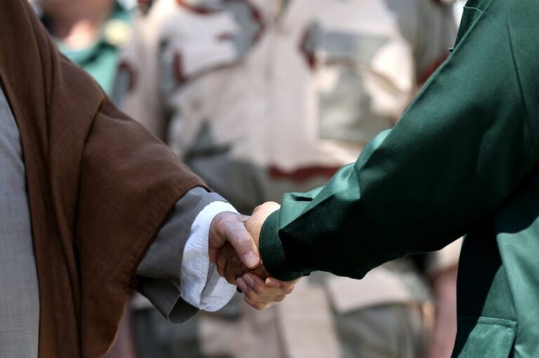 Two people are shaking hands, one in a brown robe, the other in a green uniform, suggesting formality and mutual respect. Background shows blurred uniforms.