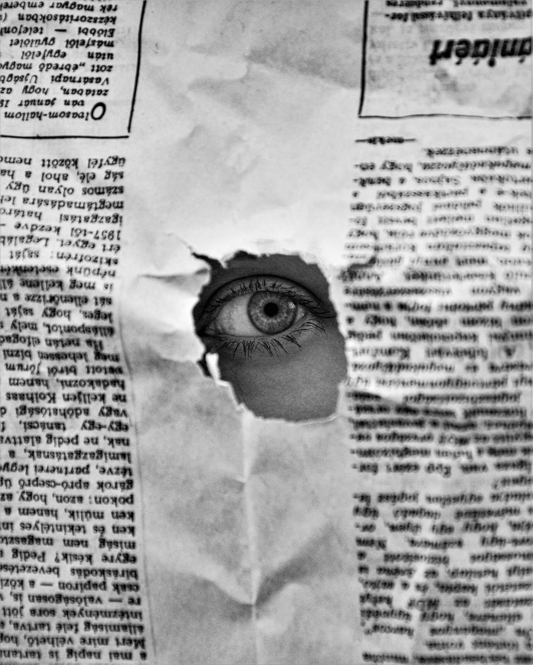 An image of an eye looking through a hole in a sheet of newspaper.