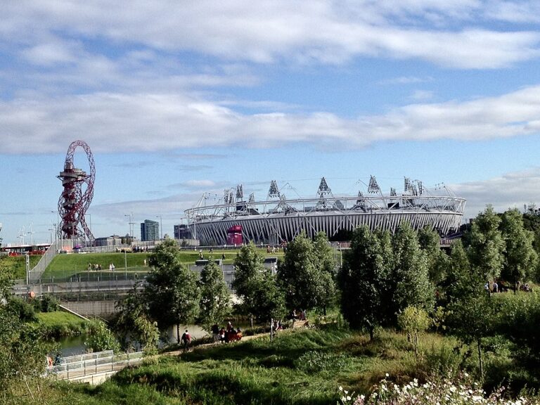 A large stadium with a white roof and angular supports sits next to a red spiral sculpture, surrounded by trees and grassy areas under a partly cloudy sky