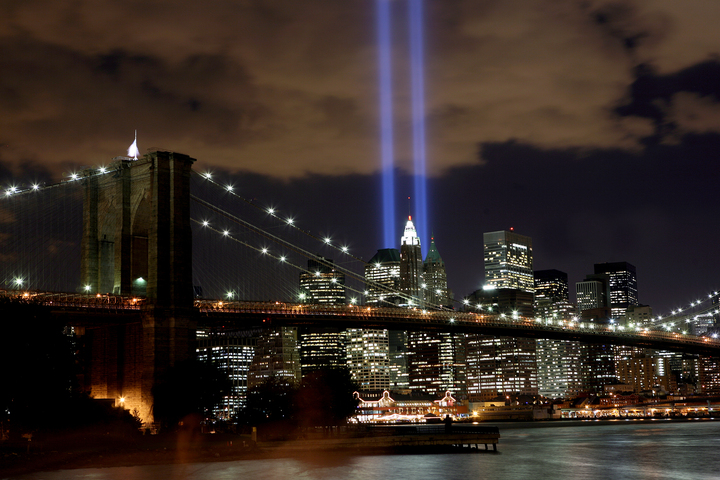 Brooklyn Bridge at night with illuminated Manhattan skyline and two blue Tribute in Light beams shining upward into the sky.