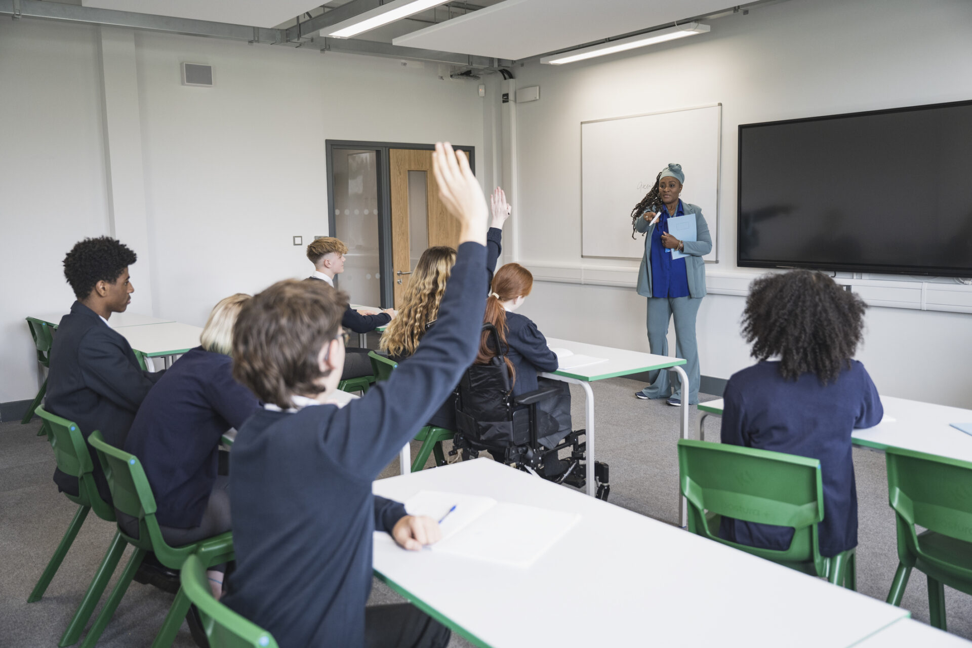A teacher stands at the front of a classroom, speaking to students seated at desks; two students have their hands raised.